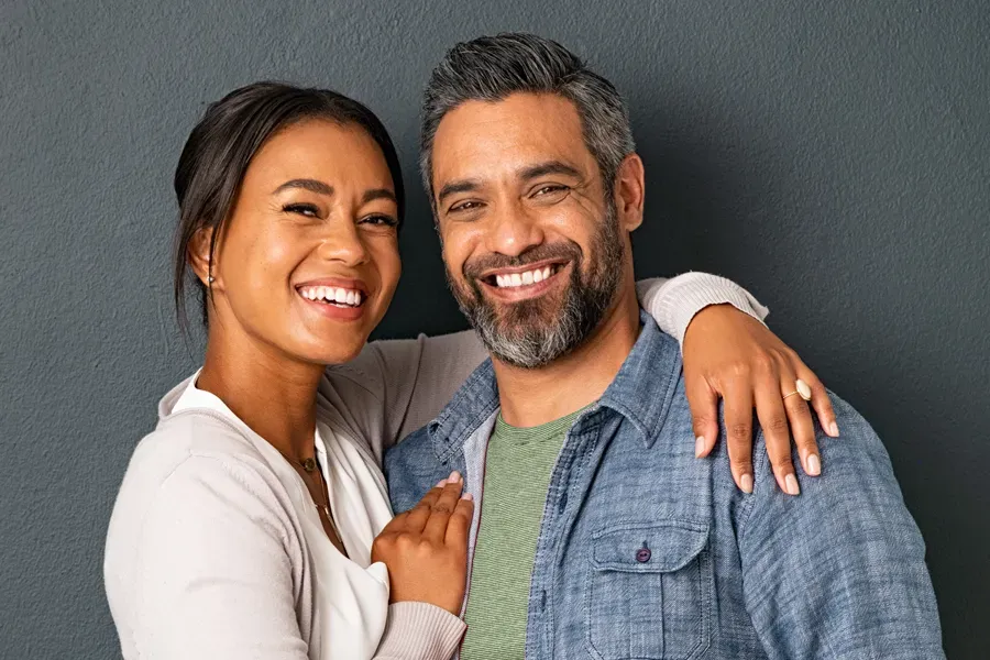 Bioidentical-Hormone-Therapy-Doctor A middle-aged couple with their arms around each other standing against a gray background and smiling, happy with the bioidentical hormone therapy they received from Marina Fay, PA-C in Aventura-North Miami.