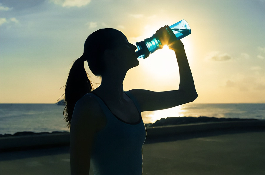 Chelation-and-Detoxification-Clinic A woman in exercise clothes drinking from a water bottle on the beach, with the water and sunset in the background. Get chelation and detoxification treatment from Marina Fay, PA-C in Aventura-North Miami.