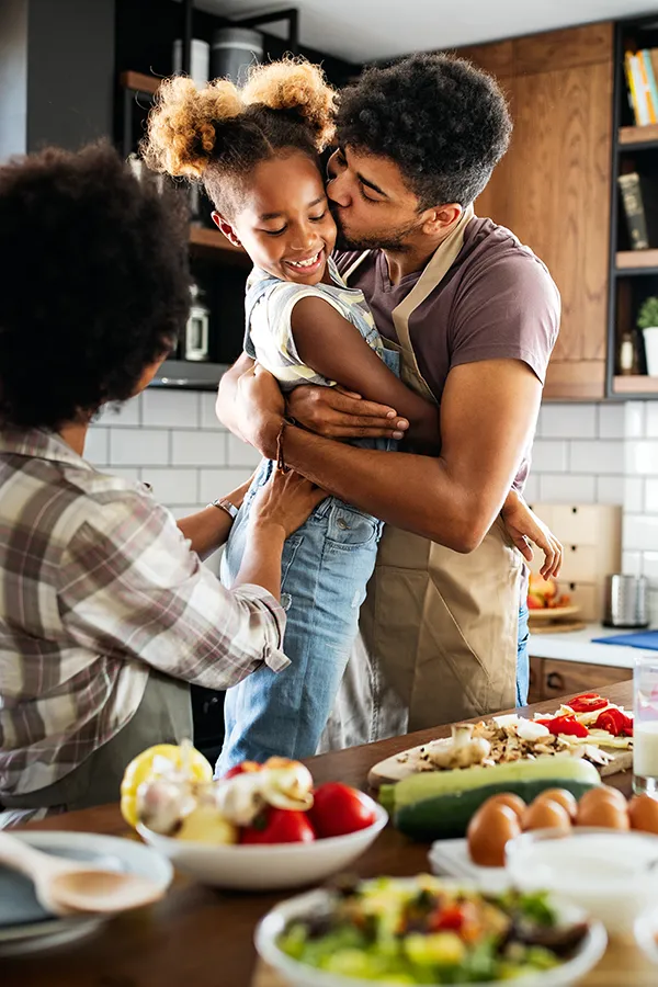 Gut-Health-Treatment A man holds his daughter and kisses her cheek as the family makes a healthy dinner in the kitchen. Get help for digestive issues and gut health from Marina Fay, PA-C in Aventura-North Miami.
