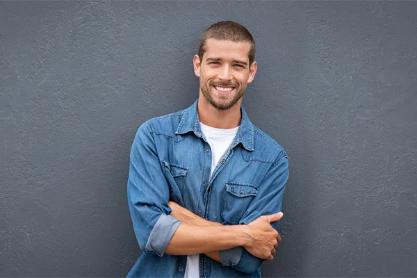 Hormones-for-Men-Doctor A man in a denim shirt stands smiling against a gray-blue wall, pleased with his testosterone hormone therapy from Marina Fay, PA-C in Aventura-North Miami.