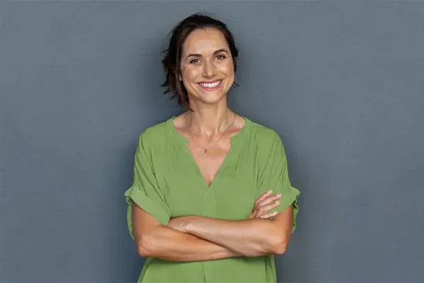 Hormones-for-Women-Doctor A woman in a green shirt stands smiling against a gray-blue wall, pleased with her hormone therapy from Marina Fay, PA-C in Aventura-North Miami.