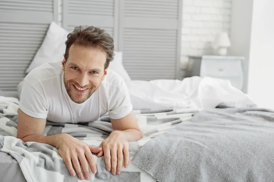 Low-Libido-in-Men-Doctor A dirty-blonde haired man lays on his stomach in bed smiling. Schedule treatment for low libido in men from Marina Fay, PA-C in Aventura-North Miami.