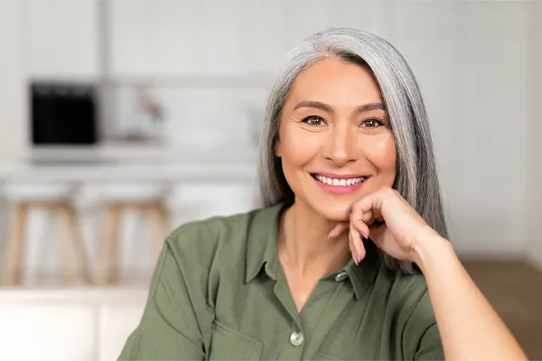 Menopause-Clinic An asian woman with long gray hair and a green shirt sitting and smiling on her couch, happy with her menopause care from [NAME} in Aventura-North Miami.