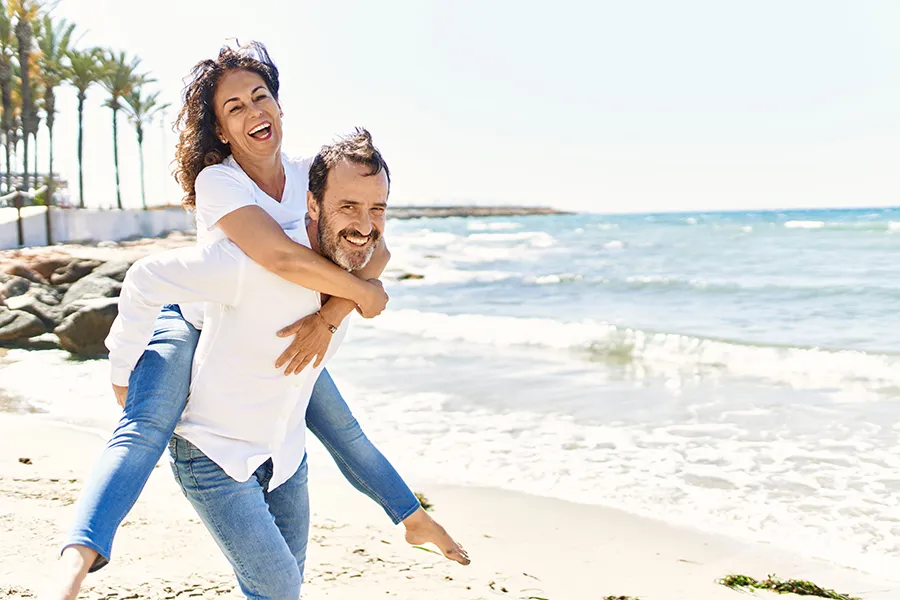 NAD-Therapy-Clinic A woman piggybacks on her husband as the middle-aged couple have fun at the beach. Get NAD Therapy from Marina Fay, PA-C in Aventura-North Miami.