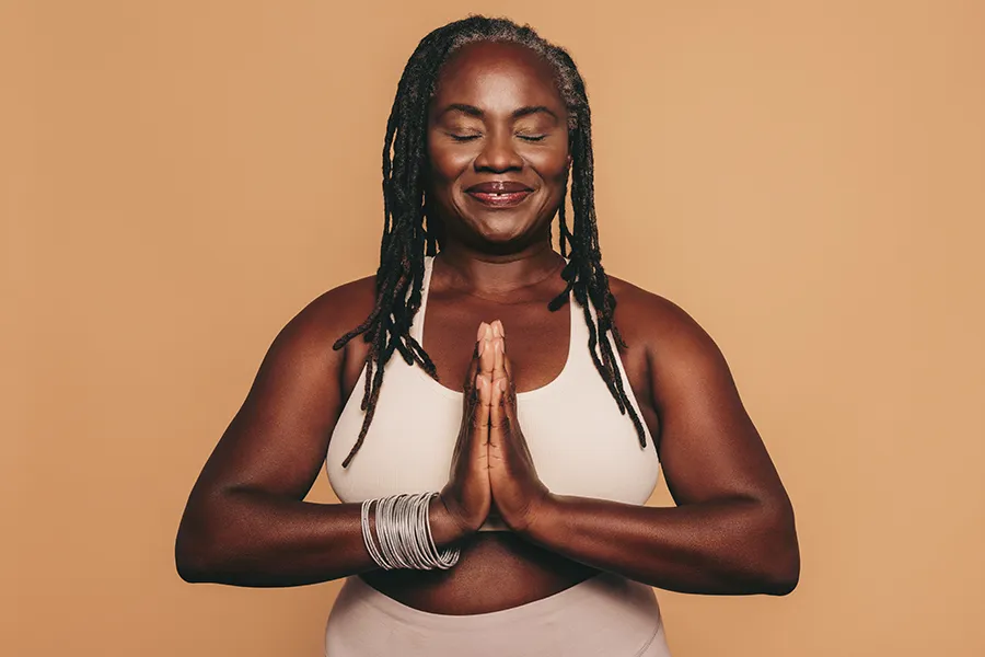 Obesity-Clinic An older African American woman confidently posing against a warm background smiling with hands in a prayer pose. Marina Fay, PA-C treats obesity using functional medicine in Aventura-North Miami.