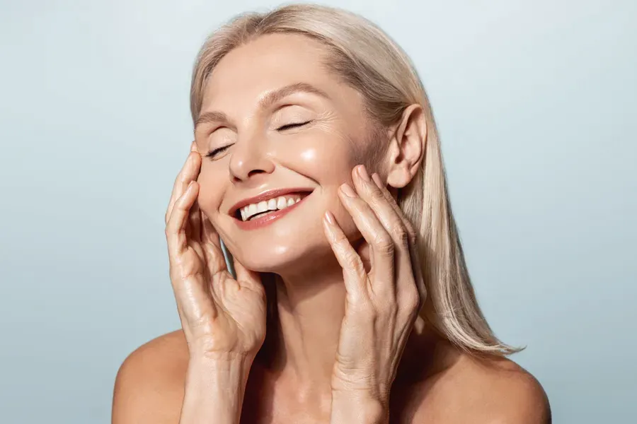PRP-Facelift-Facial-Doctor A beautiful mature blonde woman touches her face, happy with her PRP facelift and facial injections from Marina Fay, PA-C in Aventura-North Miami.