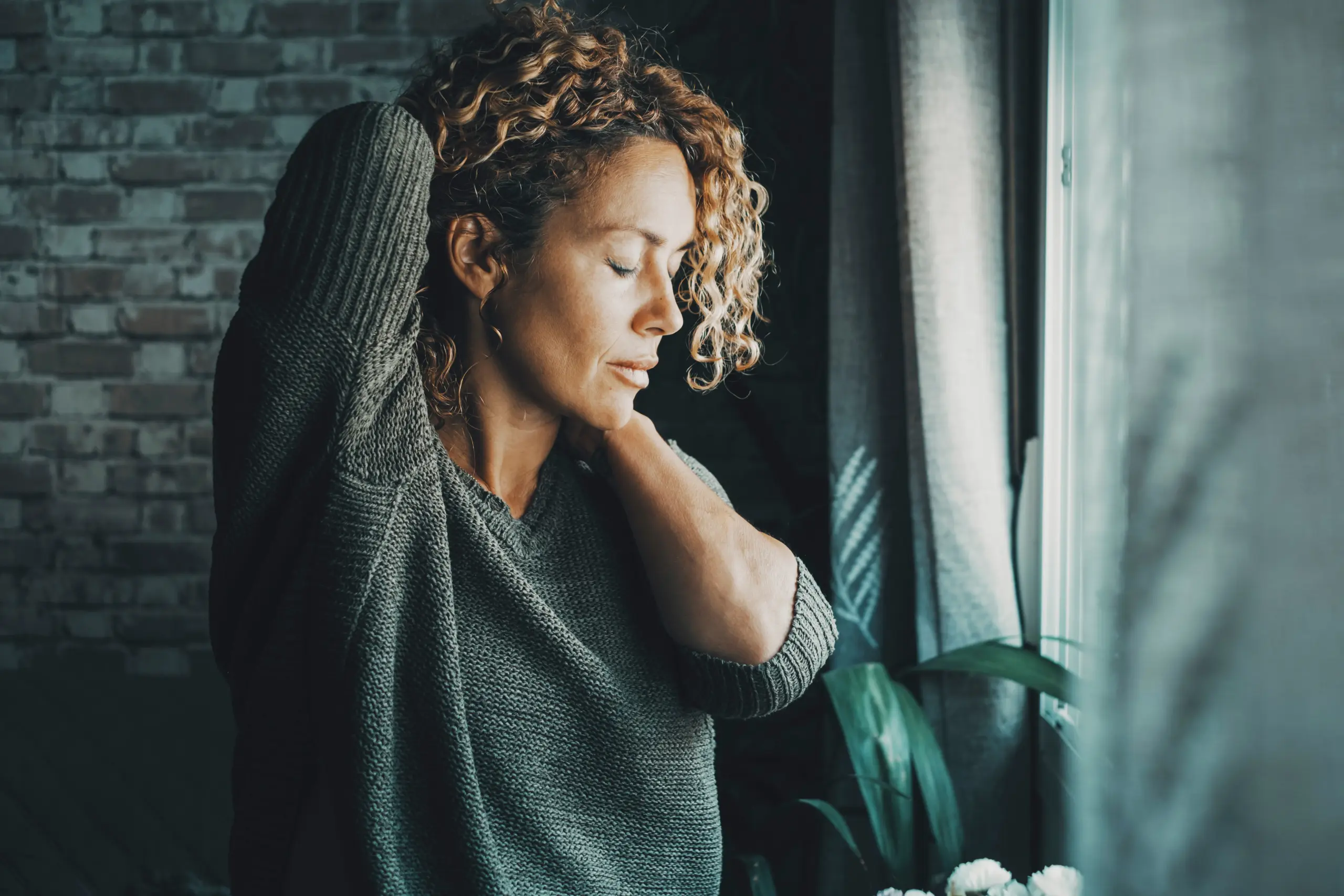 Peptide-Pain-Relief-Clinic A woman rubs her neck in pain in a dark gloomy room. Schedule peptide pain relief treatment from Marina Fay, PA-C in Aventura-North Miami.
