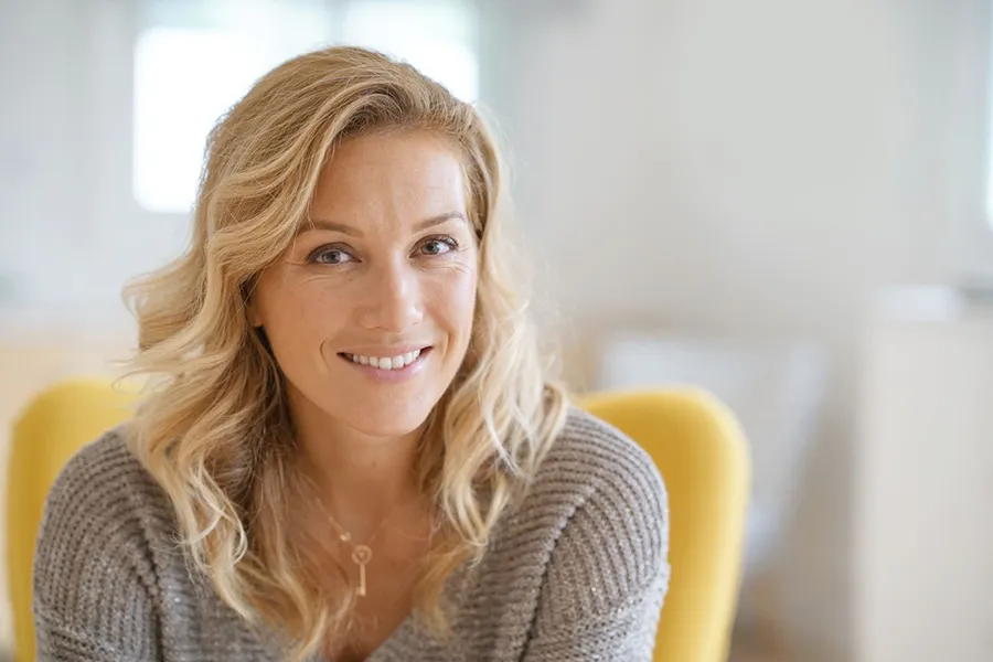 Perimenopause-Doctor A blonde woman wearing a a gray sweater sits inside in a yellow chair, smiling and happy with her perimenopause treatment from Marina Fay, PA-C in Aventura-North Miami.