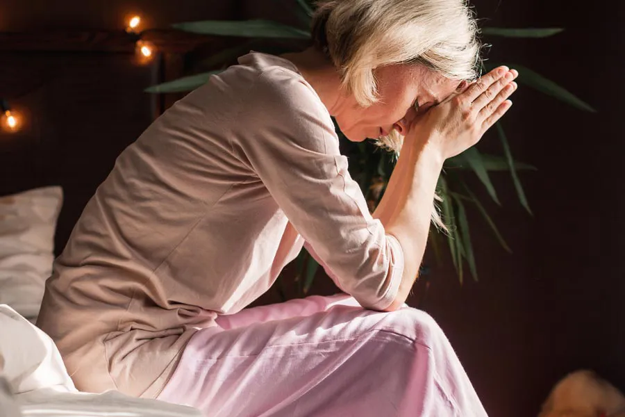 Stress-Headaches-Clinic A woman sits on the edge of her bed holding her head in her hands. Get stress headaches treatment from Marina Fay, PA-C in Aventura-North Miami.