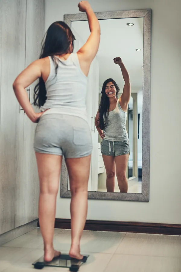 Tirzepatide-Treatment A woman in workout clothes stands on a scale looking in the mirror and pumping her hands in celebration of her successful Tirzepatide treatment from Marina Fay, PA-C in Aventura-North Miami.